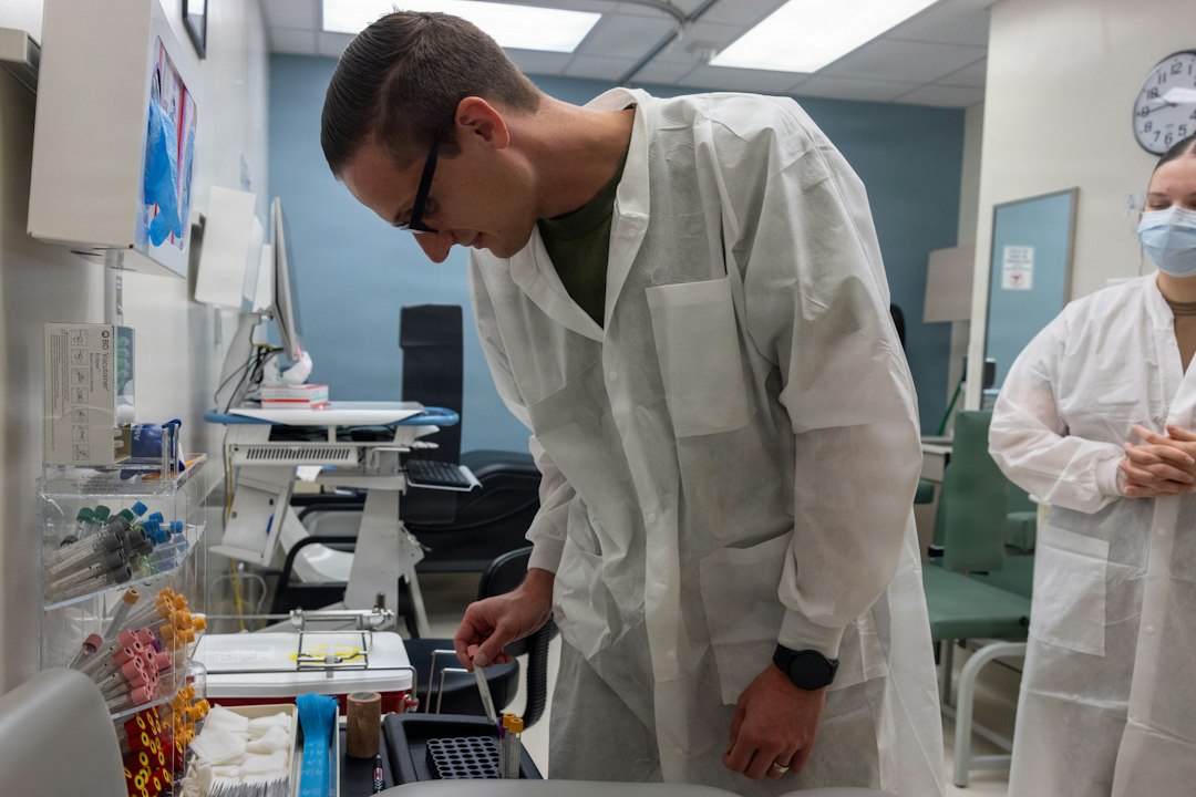 A learner in a lab coat works through a hands-on clinical training station while an instructor looks on.